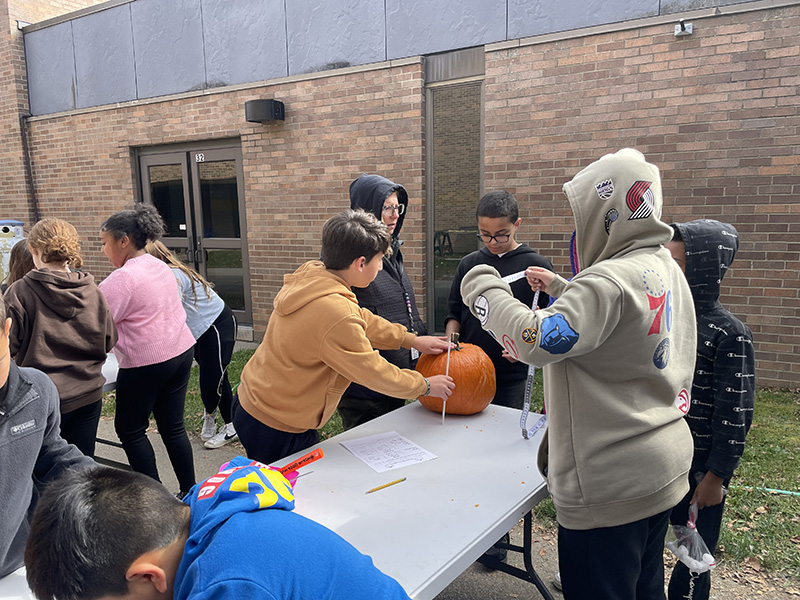 Middle school kids at a table with pumpkins. They are measuring them and taking the top off.