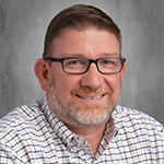 A man with short hair and a short beard smiles. He is wearing glasses and is wearing a plaid button-down shirt.