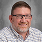 A man with short hair and a short beard smiles. He is wearing glasses and is wearing a plaid button-down shirt.