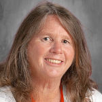 A woman with longer light hair smiles. She is wearing a white blouse.