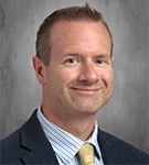 A man with short hair smiles in a professional portrait. He is wearing a dress shirt, jacket and gold tie.