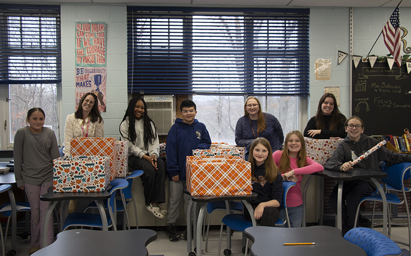 A group of seven middle schoolers and two women stand behind several boxes of food. The boxes are covered in fall-themed paper.