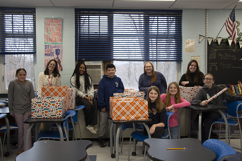 A group of seven middle schoolers and two women stand behind several boxes of food. The boxes are covered in fall-themed paper.