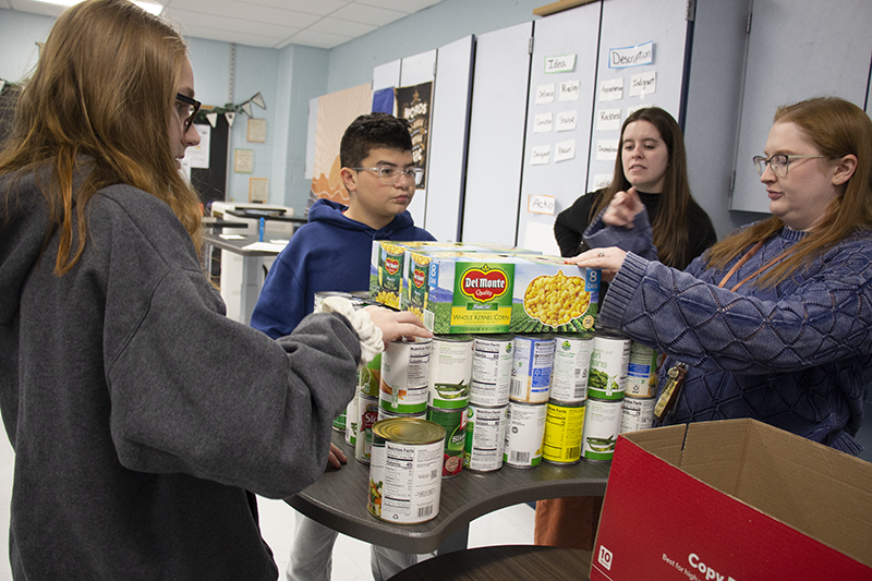 Three middle school students and a woman stand near a stack of canned vegetables.