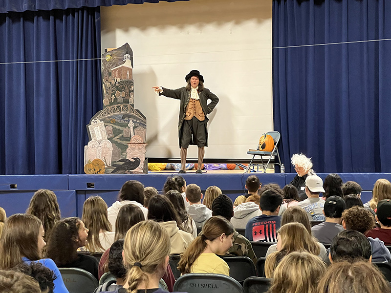 A man stands on stage dressed in 1800s period clothing, performing The Legend of Sleepy Hollow. A large group of kids is in front of him watching.