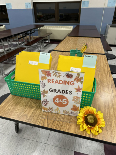 A green basket of yellow folders sit on a table. There is a sign that says Reading Grades 4-5 and there is a sunflower on the table.