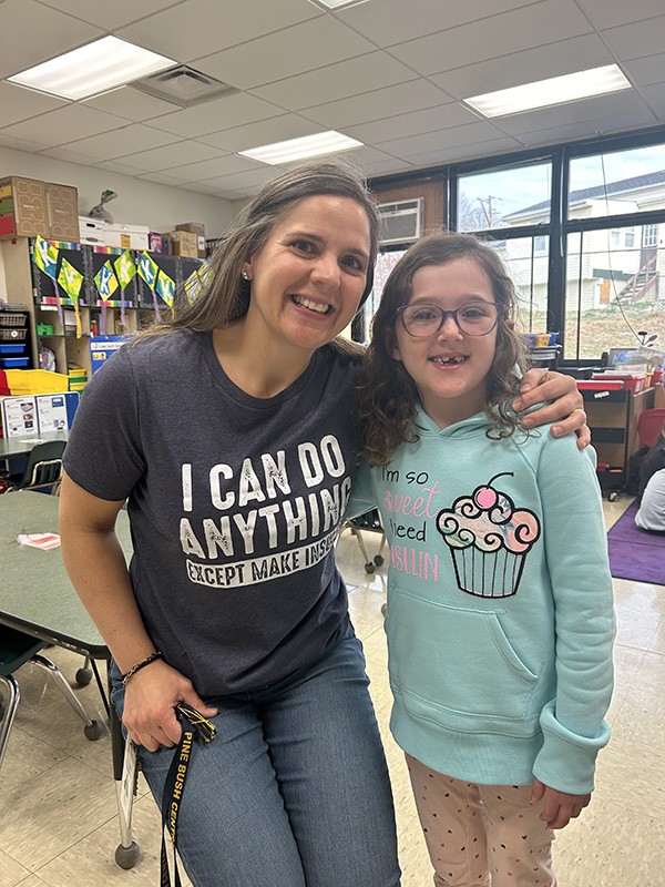 A woman with long light hair smiles and puts her arm around a little girl. They are both smiling. The woman is waring a shirt that says I can Do Anything (except make sure).