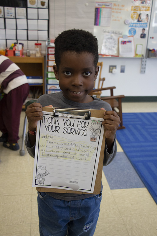 A first grade boy smiles as he holds up the letter that he wrote to his dad, a veteran.