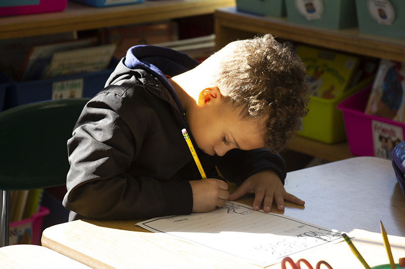 A first grade boy sits at a desk writing with a pencil.