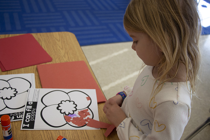 A first-grade girl with shoulder-length blonde hair pastes red pieces of paper onto a piece of paper to make a poppy.