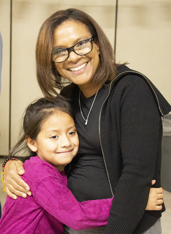 A woman with shoulder length hair and glasses hugs a little girl with long dark hair, wearing a purple sweater. Both are smiling.