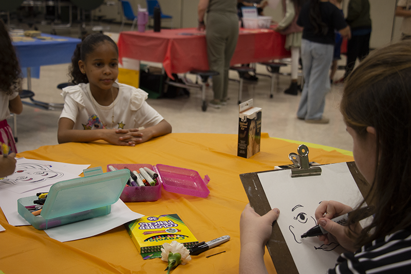 A high school girl is drawing a caricature of a little girl who is sitting across the table from her.