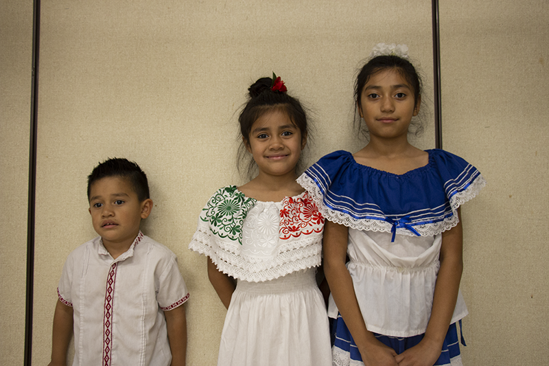 Two sisters smile standing next to their brother. The girls are wearing colorful dresses.