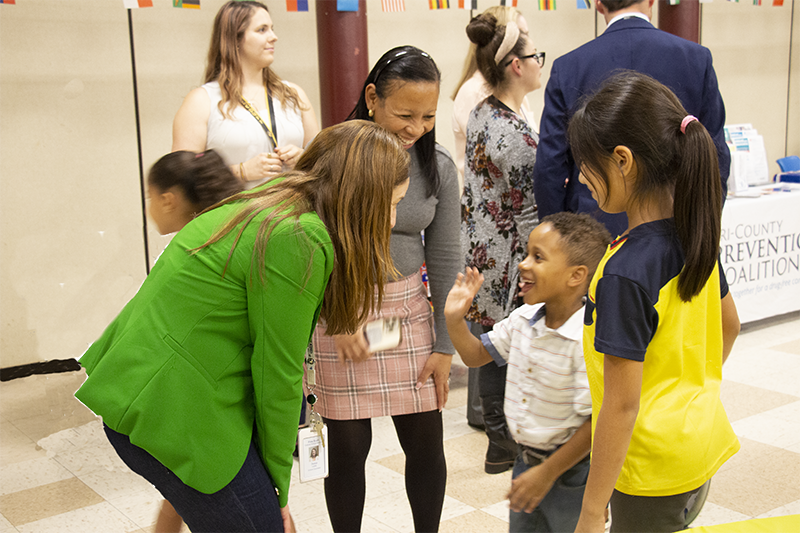 A woman with long reddish hair bends down to talk to a little boy who is smiling and waving. Another woman stands nearby smiling.