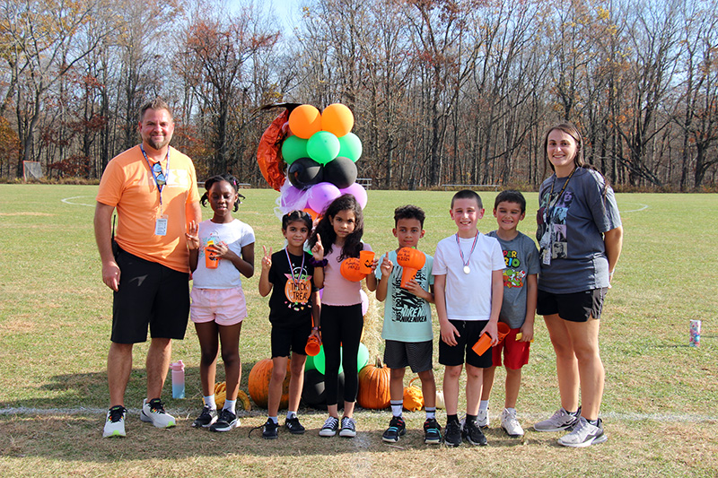 Six third-grade students - three boys and three girls - stand together in front of a column of balloons. Some are holding pumpkins. A man is on the left and a womanis on the right.