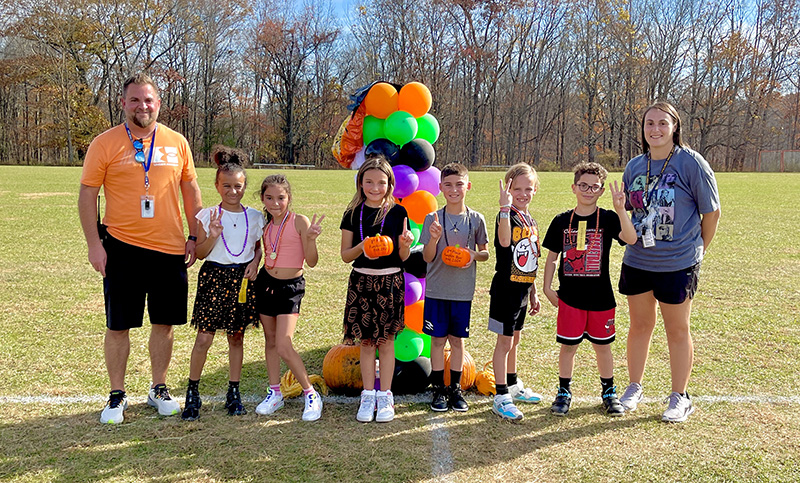 Six fourth-grade students stand in front of a bunch of balloons. On either side of them is an adult.