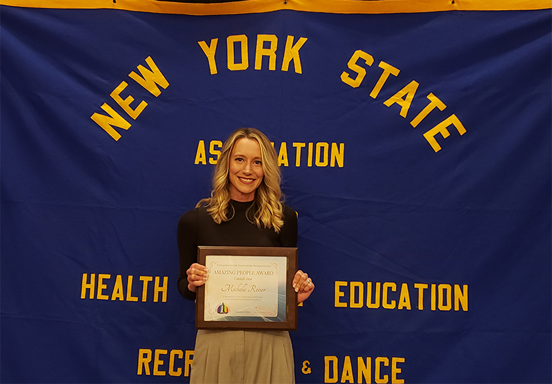 A woman with long blonde hair smiles and holds a certificate. Behind her is a large banner that says New york State Association of Health, Physical Education, Recreation and Dane.