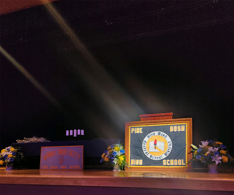 A stage with a podium that has a large sign that says Pine Bush High School. There is a beam of light shining on it.