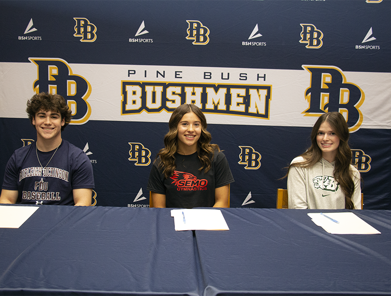 Three high school seniors - a boy at left, and two girls at center and right, sit at a table and smile. Behind them is a banner that says PB Pine Bush Bushmen in blue and gold.