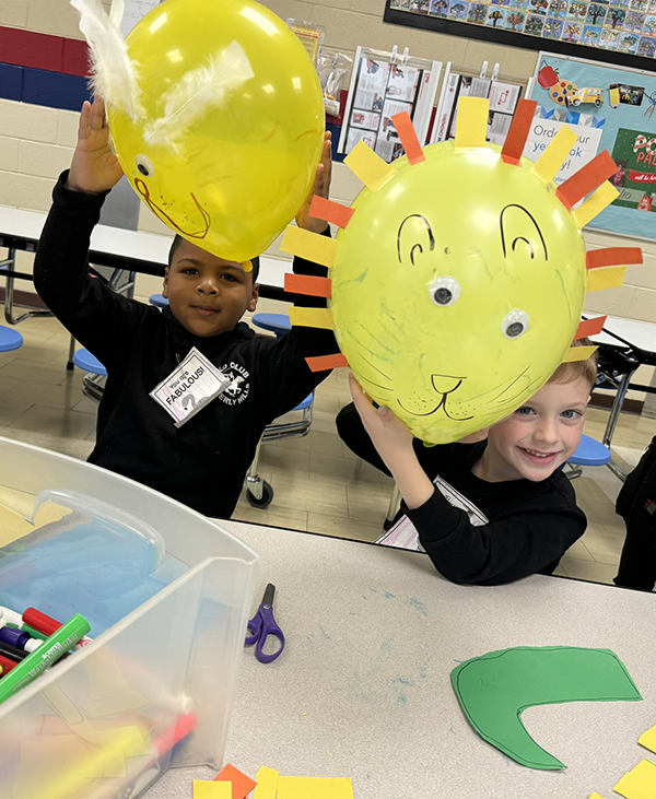 Two kindergarten boys holding up large yellow balloons they have decorated. They are smiling.