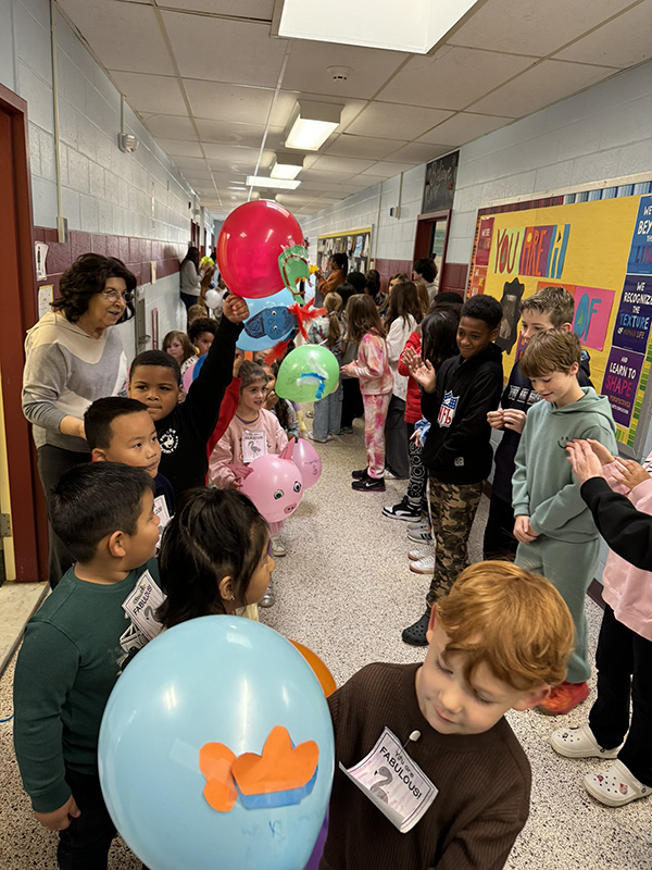 A large group of elementary school kids gather in a hallway with a path open in the center for a parade.