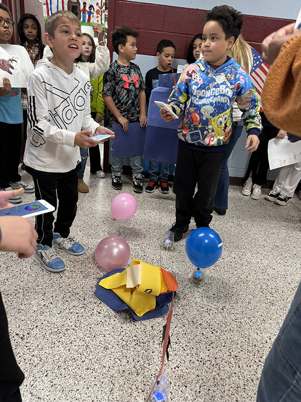 An older elementary age kid uses Spheros, a robotics device, to roll his balloon down the hall.