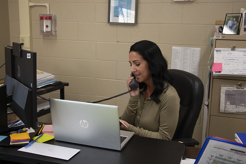 A woman with long dark hair sits at a desk with a computer reading a book aloud. She is holding a phone.