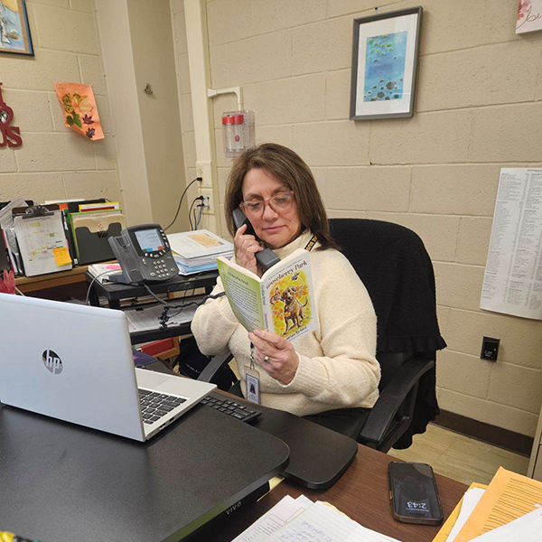 A woman with long dark hair sits at a computer. She is reading a book and holding a phone in her hand.