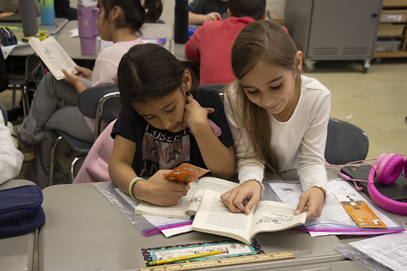 Two fourth-grade girls sit at a desk reading a book together.