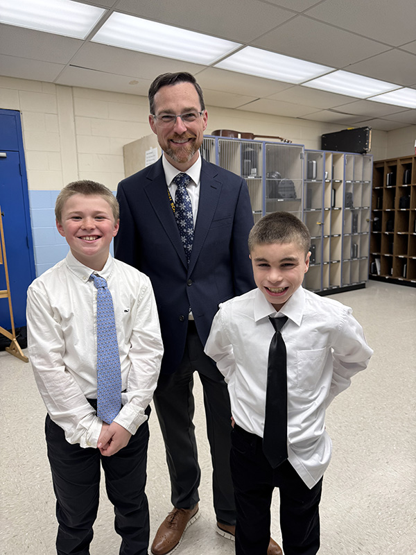 A man with short dark hair, wearing glasses and a suite and ti smiles as he stands with two older elementary boys, both wearing white shirts and ties. They are smiling too.