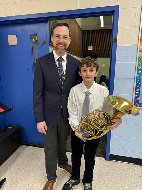 A man with short dark hair, glasses and a suit and tie smiles while standing with a young man wearing a white shirt, gray tie and holding a French horn.