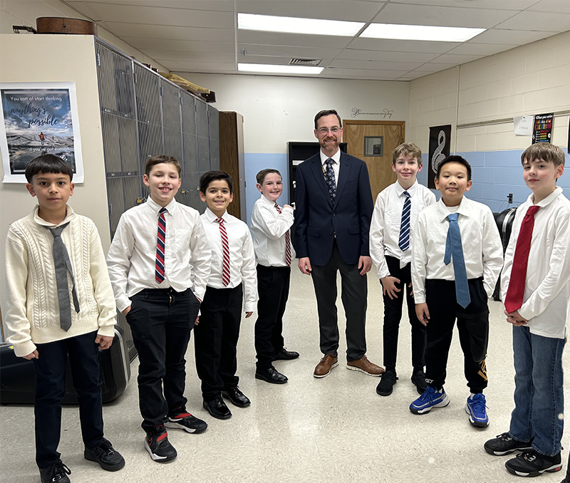 A man with dark hair, wearing glasses and a suit and tie smiles as he stands with seven older elementary students, all wearing shirts and ties.