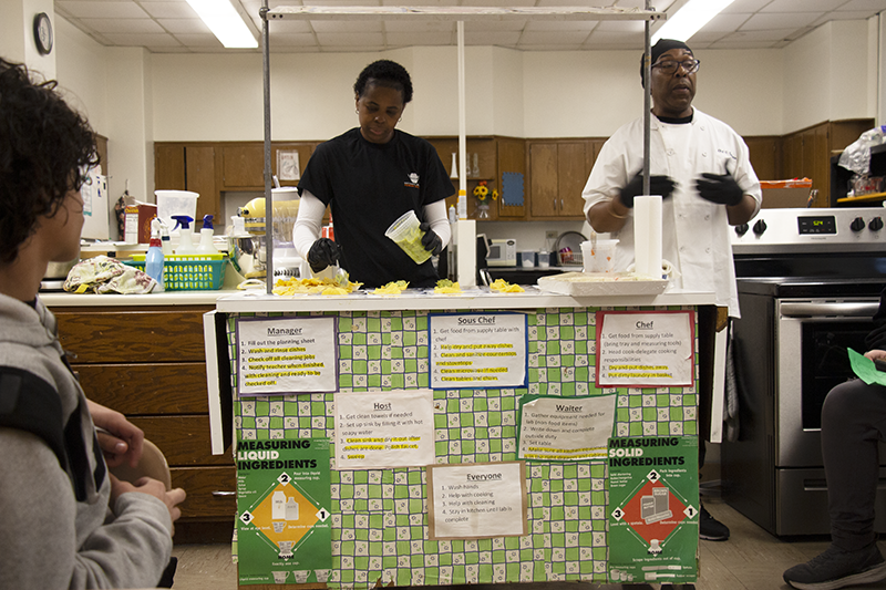 A man in a white shirt and a person next to him wearing a black shirt put together salsa and guacamole. On the front of the table, are papers that have job titles in the restaurant industry and their duties.