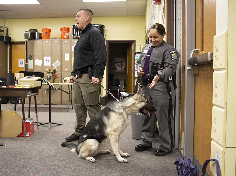 Two state troopers talk to a group of middle school students. One is holding a leash with a large dog at the end.