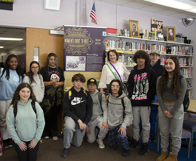 A group of 10 middle schoolers stand with a woman who is dressed in a high neck white blouse and long skirt with a sash across her body showing she is a suffragette.