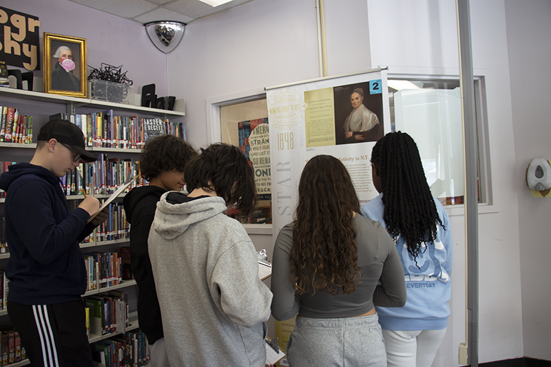 Four eighth-grade students write on their clipboards as they look at a large display of historical information.