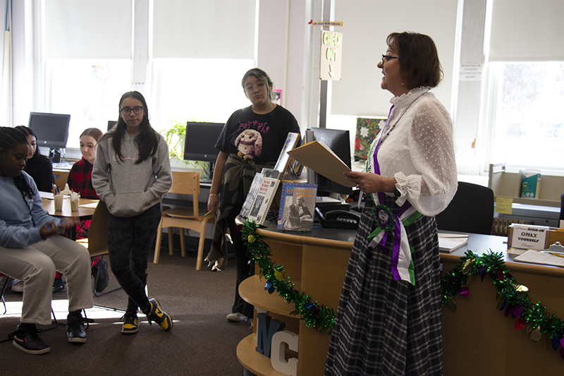 a woman in a long-sleeve white blouse and long skirt talks to a group of eighth-grade students  in a library.