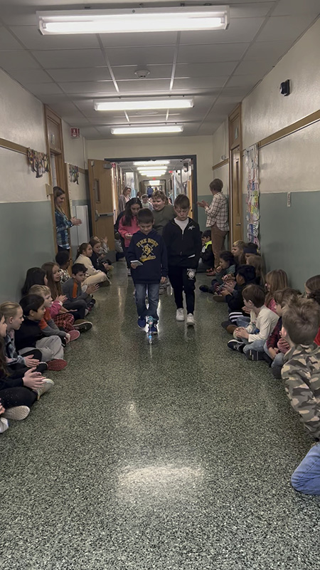 A hallway is lined with students sitting against a wall and kids are coming down the hall with small balloons riding on a spheros  they are controlling from their device.