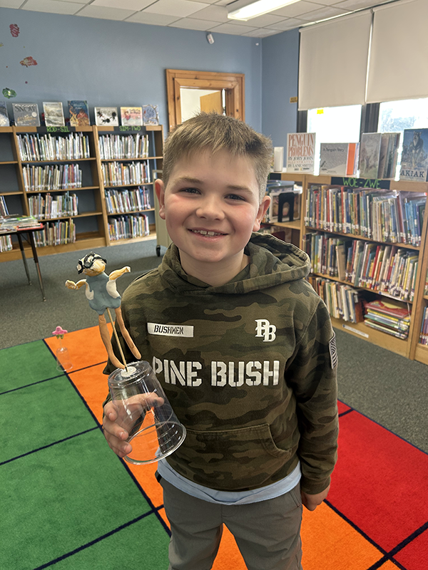 A fourth-grade boy with short light hair smiles. He is holding a little clay balloon he made.