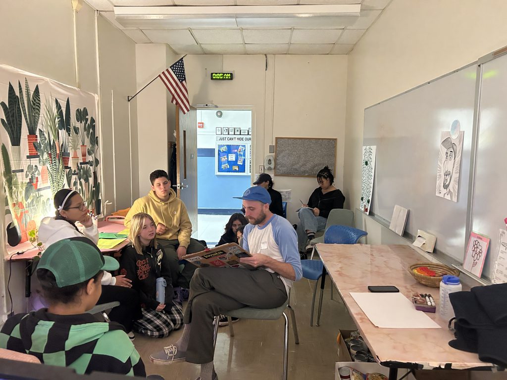 A man sits in the center of a group of middle school kids drawing on white paper.