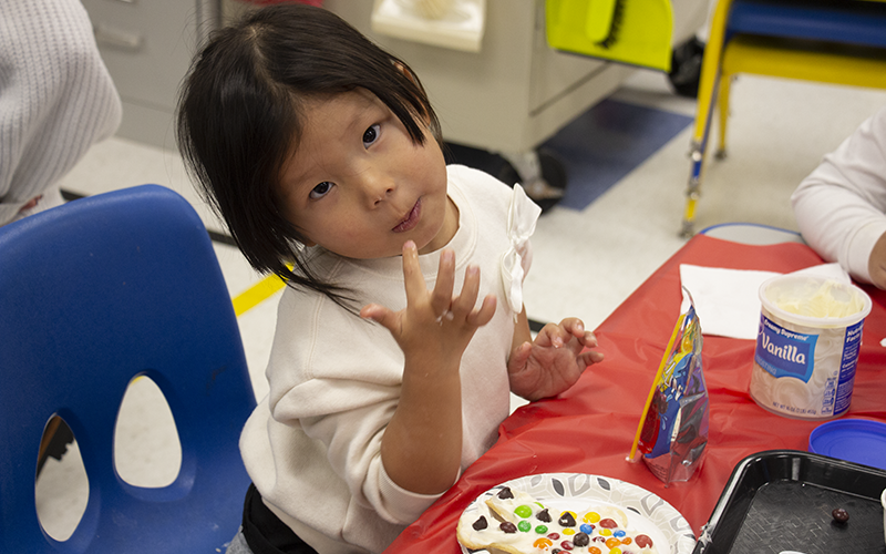 A pre-K girl with short dark hair tilts her head to the side whole licking frosting off her fingers. On the plate in front of her is a cookie with frosting and candies on it.