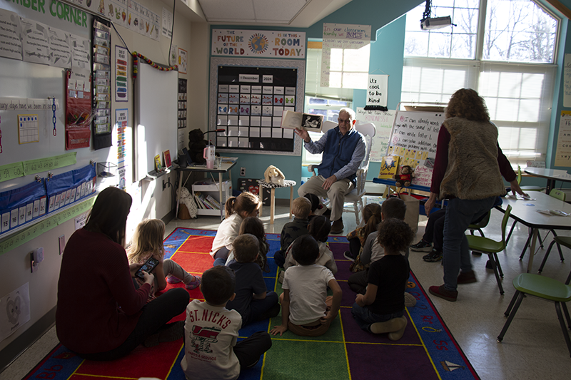 From the back of a classroom, a class of kindergarten kids sit on a multi-colored rug listening to a man read a book to them.