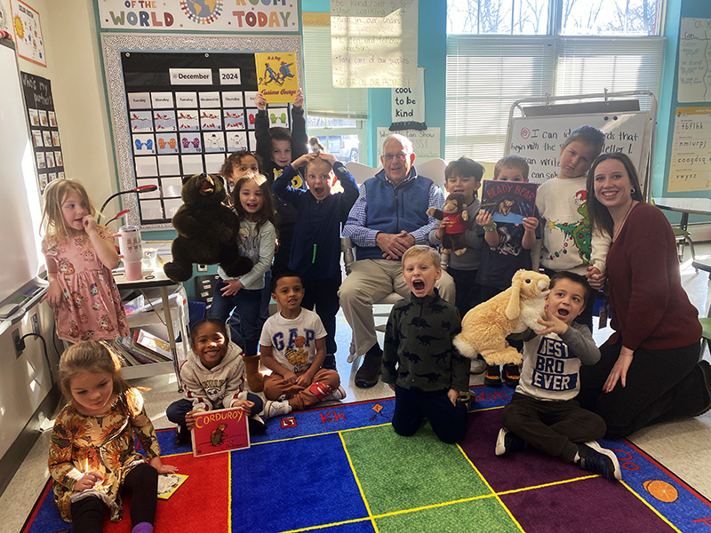 A group of 13 kindergarten kids smile, with some holding up puppets. There are two adults in the picture too.