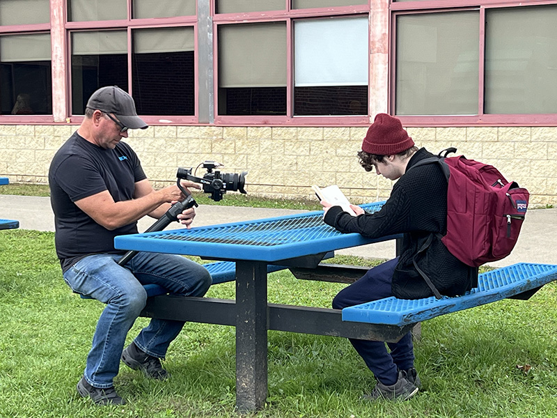 A high school student wearing a backpack and red hat, sits at a blue picnic table reading a book. A man wearing jeans and a black shirt and a baseball cap holds a camera while he films the kid.