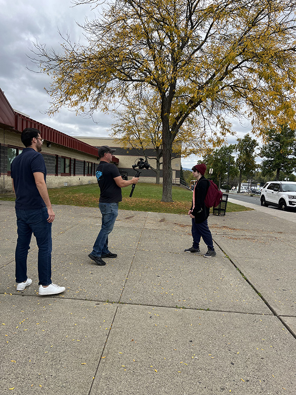 A man watches as a high school boy wearing a backpack and a red hat is filmed as he walks into his school.