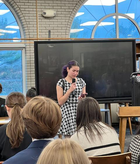 A high school girl in a blue and white plaid dress stands in front of a group of other high school kids speaking.