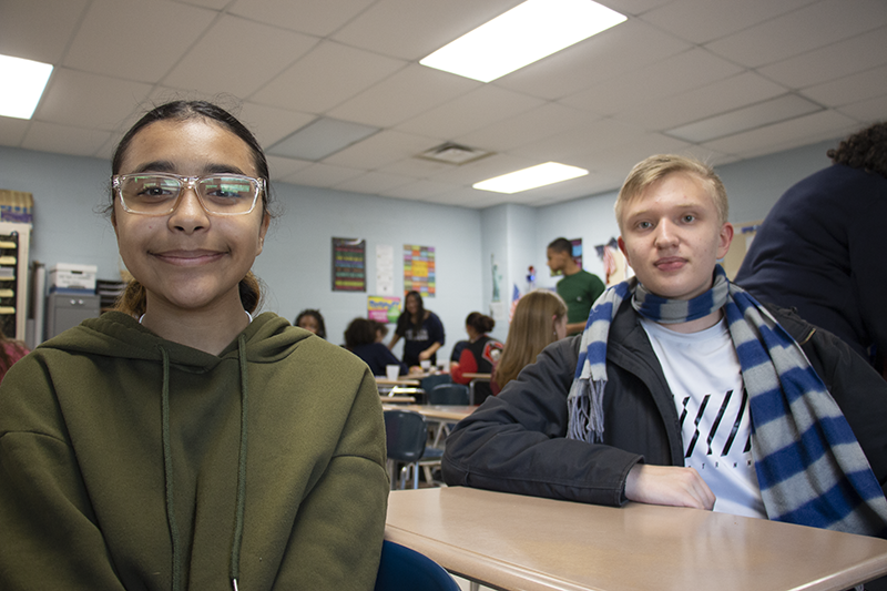 A young woman on the left, waring glasses and a gree hoodie, and a high school boy on the right with short blonde hair wearing a light blue shirt and a plaid scarf.