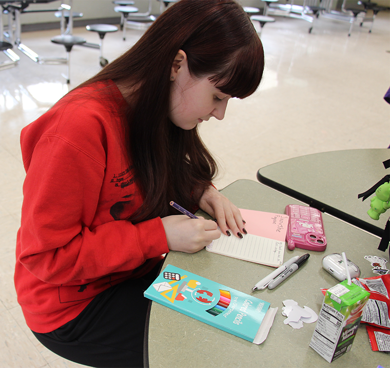 A high school girl with long dark hair wearing a red hoodie writes in a journal on a table.