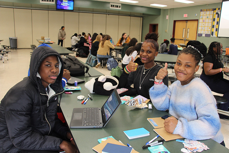 Three high school students sit at a table journaling. Two on the right are giving thumbs up.