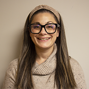 A woman with long dark hair and glasses smiles. She is wearing a tan sweater.